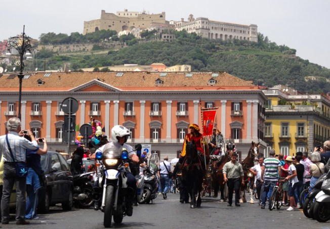 The parade of carriages leaves Piazza del Plebiscito in Naples, Italy, beneath the watchful Castel Sant'Elmo and the Certosa di San Martino